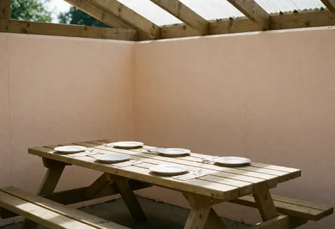 Table en bois pour pique-nique sous un toit, dressée avec assiettes et couverts prêts à être utilisés, dans un espace aux murs beiges éclairé par la lumière naturelle.