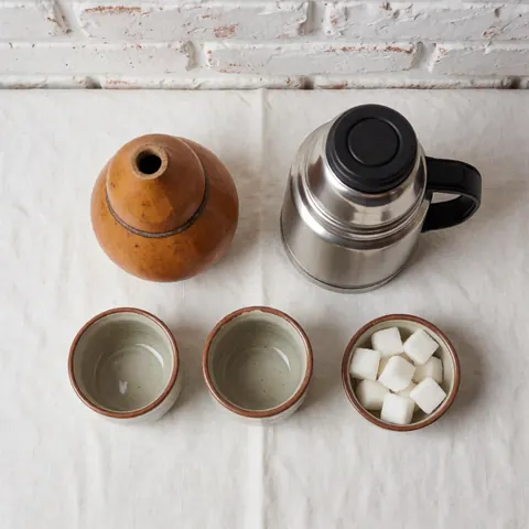 Service à maté complet avec gourde, deux tasses, thermos et sucre dans un bol, posé sur un tissu blanc devant un mur de briques.