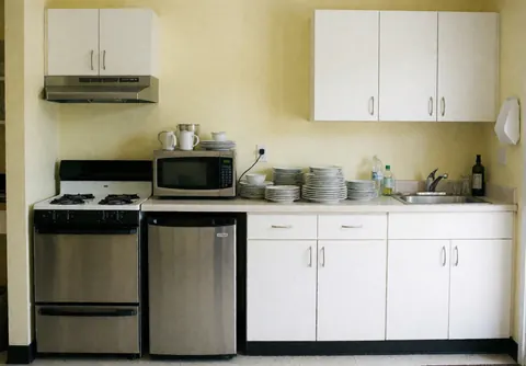 Cuisine compacte d'appartement avec cuisinière, micro-ondes, réfrigérateur et évier, murs jaunes clairs, ustensiles et assiettes rangés.