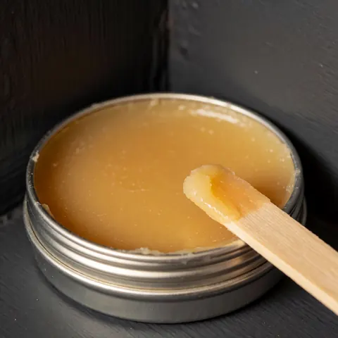 Boîte métallique contenant de la cire de carnauba jaune accompagnée d'un applicateur en bois posé sur un mur gris foncé sous un éclairage naturel et chaleureux.