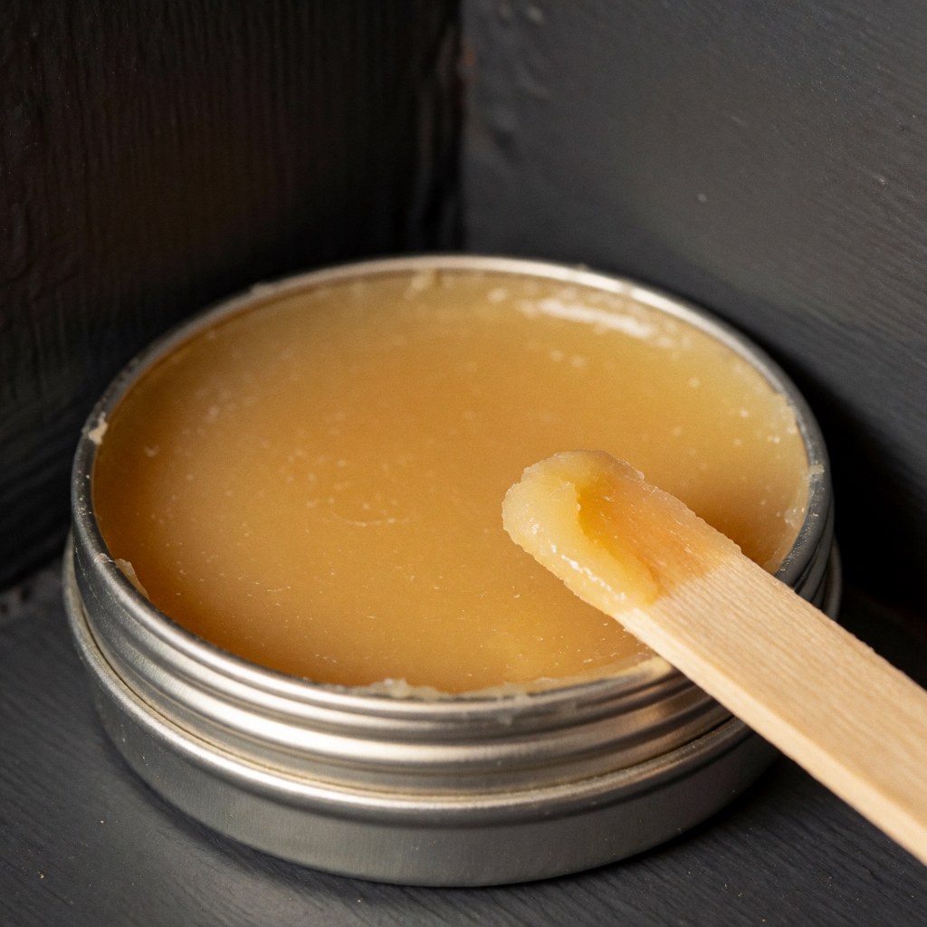 Boîte métallique contenant de la cire de carnauba jaune accompagnée d'un applicateur en bois posé sur un mur gris foncé sous un éclairage naturel et chaleureux.