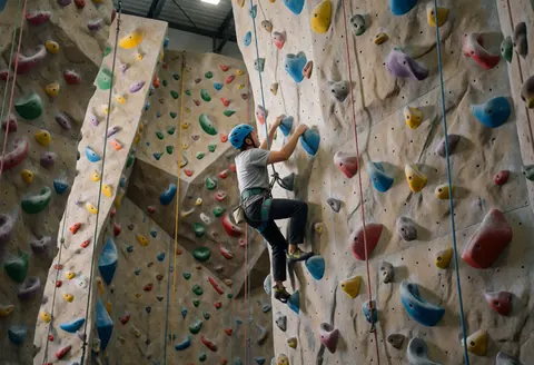 Escaladeur progressant sur un mur texturé aux prises colorées dans un gymnase intérieur, équipé d'un casque et d'une baudruche, sous un éclairage professionnel.