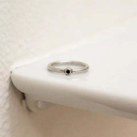 Bague en titane ornée d'une perle noire posée sur une surface blanche, texture métallique visible et éclairage neutre pour présentation de bijoux corporels.