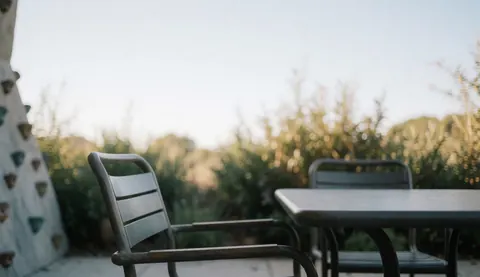 Table et chaises en métal sur une terrasse extérieure, avec un mur d'escalade flou à gauche et de la végétation naturelle en arrière-plan, éclairé par une lumière douce du soleil.