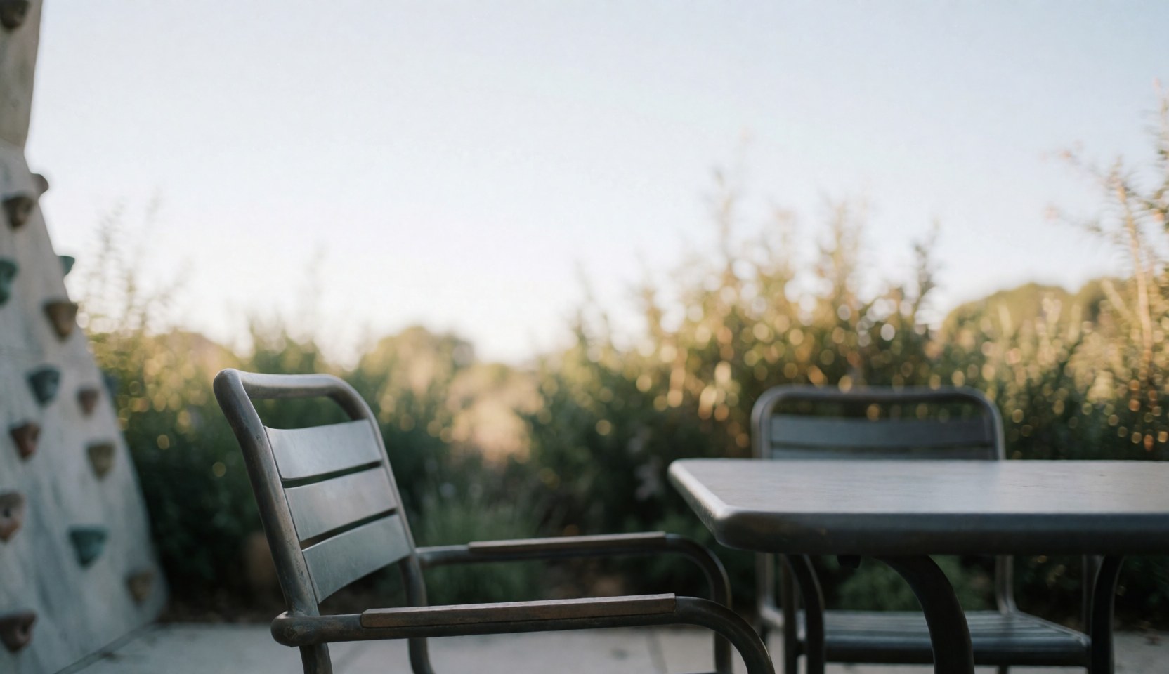 Table et chaises en métal sur une terrasse extérieure, avec un mur d'escalade flou à gauche et de la végétation naturelle en arrière-plan, éclairé par une lumière douce du soleil.