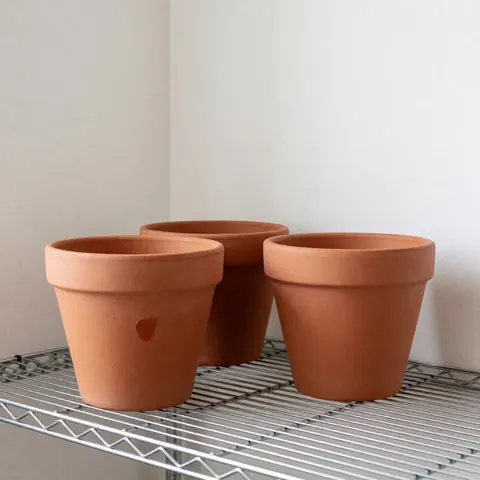 Trois pots en terre cuite non peints posés sur une étagère métallique, avec des murs blancs et un éclairage doux du matin mettant en valeur la texture naturelle.