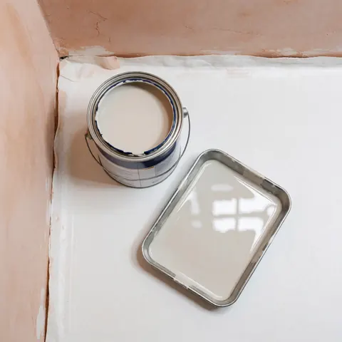 Vue de dessus d'un pot de peinture synthétique brillante et d'un bac posés sur une toile blanche, avec des murs en plâtre clair en arrière-plan, évoquant la préparation d'une peinture intérieure.
