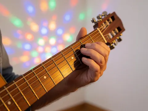 Gros plan sur une main touchant les cordes d'une guitare acoustique, éclairée par des lumières colorées avec un effet bokeh sur fond blanc.