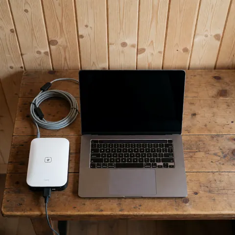 Un ordinateur portable connecté à un modem satellite repose sur une table en bois rustique, illustrant l'accès internet dans un refuge de montagne.