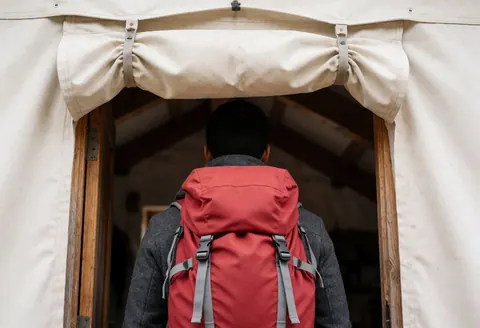 Un randonneur portant un sac à dos rouge et une veste grise franchit l'entrée d'un refuge de montagne doté d'une bâche beige et d'une structure en bois, révélant un intérieur aux poutres apparentes.