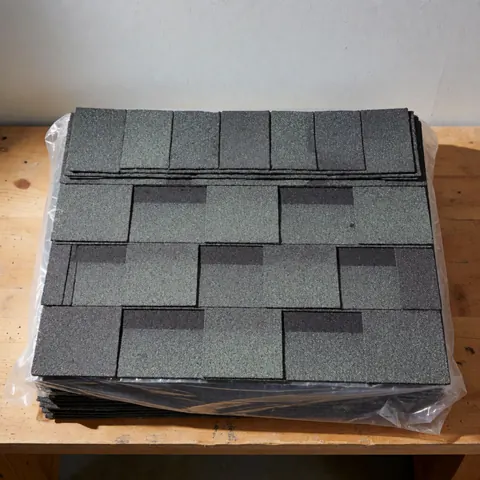Paquet de tuiles d'asphalte grises à texture granulaire, emballées dans du plastique, posé sur un banc en bois avec un mur en arrière-plan.