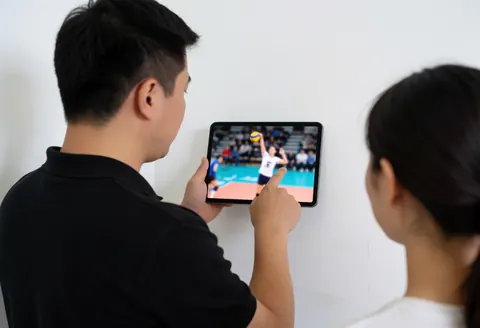Un entraîneur de volleyball examine attentivement des images tactiques sur une tablette numérique dans un environnement professionnel.