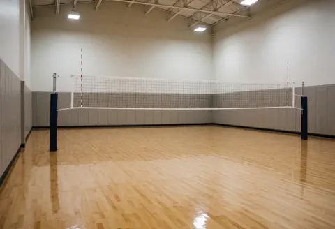 Terrain de volleyball vide au sol en bois brillant, filet blanc sur poteaux noirs, murs avec panneaux de sécurité et éclairage plafond.