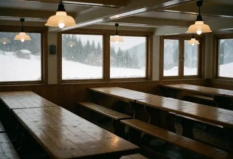 Intérieur d'un refuge de montagne vide avec tables rustiques, bancs, suspensions et fenêtres montrant la neige tombante. Ambiance chaleureuse.