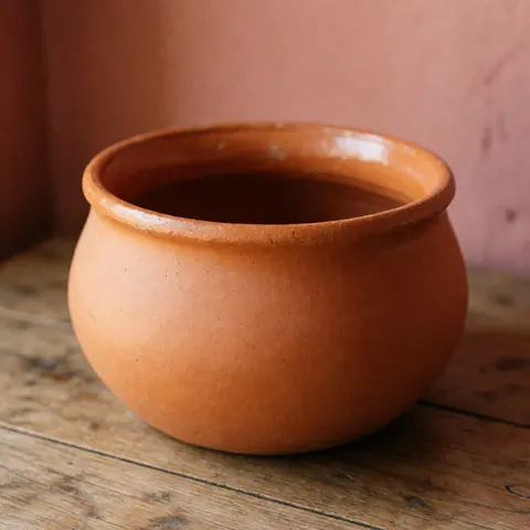 Cocotte artisanale en terre cuite mate posée sur une table en bois rustique, fond rose pâle et lumière naturelle mettant en valeur la texture de l'argile.