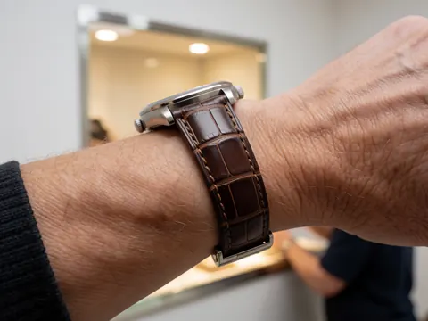 Gros plan sur la fermeture d'une montre avec un bracelet en cuir de crocodile, sur fond de vitrine éclairée. Détail de la texture du cuir et du fini métallique.