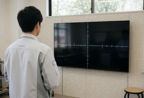 Un technicien en uniforme blanc examine un écran de télévision mural de soixante-cinq pouces affichant une croix blanche, dans un environnement ordonné et éclairé par la lumière naturelle.