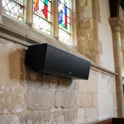 Enceinte linéaire noire fixée sur un mur en pierre dans une église, éclairée par la lumière naturelle filtrant à travers les vitraux colorés.