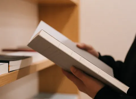 Gros plan d'une épingle fixée sur la couverture d'un cahier bilingue, tenu par des mains dans une papeterie avec des étagères en bois.