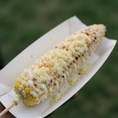 Épi de maïs grillé garni de mayonnaise blanche, de fromage râpé et d'épices, posé sur du papier blanc devant un fond vert.