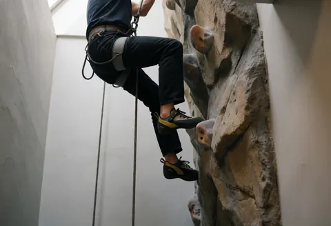 Personne escaladant un mur artificiel avec des chaussures, une corde et un harnais sur un mur en pierre texturée sous la lumière naturelle.