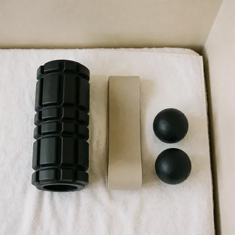 Outils de récupération sportive : un cylindre noir, une bande de résistance beige et des balles de massage posés sur une serviette blanche dans un environnement d'entraînement.