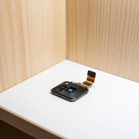 Module de caméra de smartphone avec capteur et câble flex sur fond blanc, murs en bois clair, éclairage professionnel pour réparation.