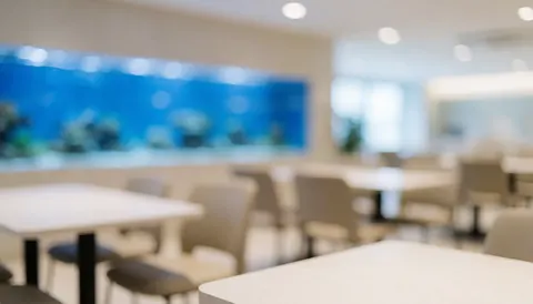 Arrière-plan d'une cafétéria moderne avec un grand aquarium bleu sur le mur, des tables et chaises floues en premier plan, ambiance lumineuse et aérée.