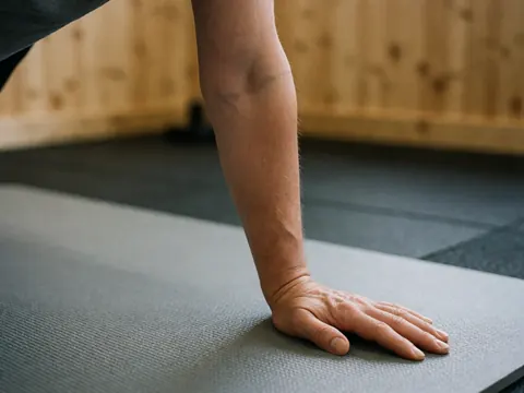 Gros plan d'une main adulte reposant sur un tapis de fitness avec un mur en bois et un sol flou en arrière-plan.