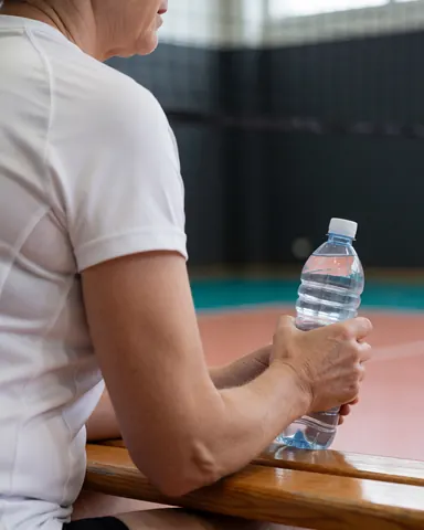 Un athlète âgé se repose sur un banc en bois près d'un terrain de volleyball, tenant une bouteille d'eau, avec un fond flou et un éclairage doux.