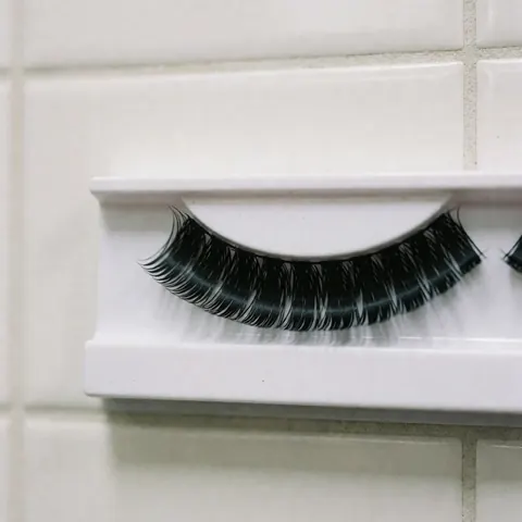 Des faux cils noirs et épais posés sur un support blanc devant un mur de carreaux blancs, avec une lumière douce idéale pour la vente de produits de beauté.