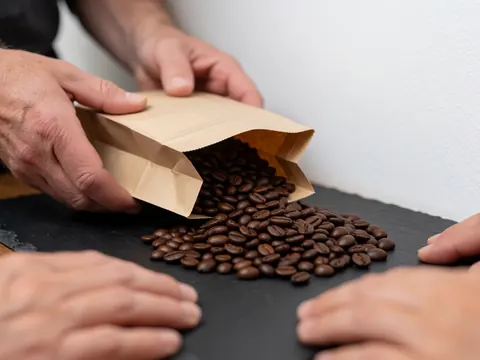Des mains versent des grains de café d'une sacoche en papier sur un plan de travail en pierre foncée, mettant en valeur la texture détaillée des grains.