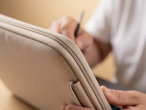 Gros plan sur une fermeture éclair métallique et la couture d'une housse beige pour ordinateur, mains effectuant un travail de précision sur fond doux et chaleureux.