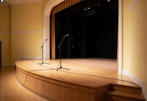 Escene courbe en bois équipée de deux microphones, câbles, rideaux noirs, murs jaunes et éclairage chaleureux prêt pour des événements.
