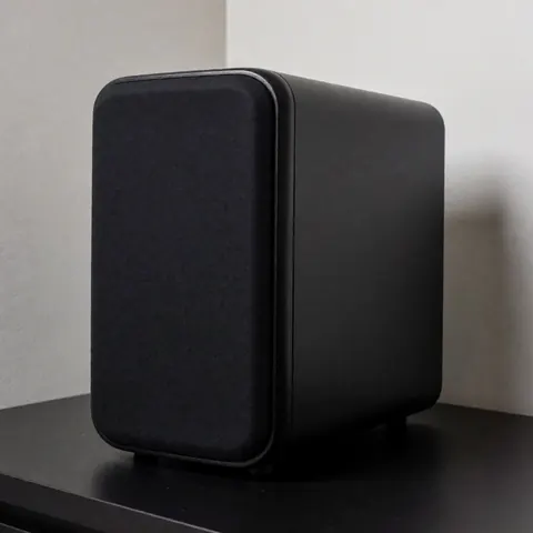 Subwoofer noir à finition mate posé sur une table noire avec des murs gris clair en arrière-plan et un éclairage doux et professionnel.