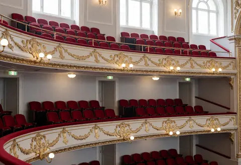 Balcons de théâtre vides avec des sièges rouges, des garde-corps dorés et des murs gris, éclairage naturel par des fenêtres hautes, idéal pour promouvoir des événements culturels.