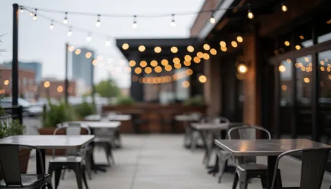 Terrasse extérieure équipée de tables et chaises en métal, éclairée par des suspensions lumineuses sur fond flou d'édifices urbains.