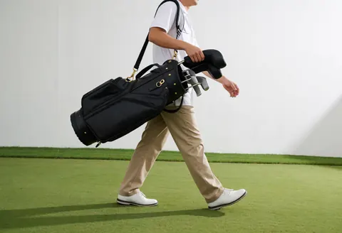 Un caddie professionnel en uniforme blanc et pantalon kaki marche avec son sac de golf sous la lumière du matin.
