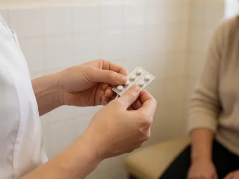 Praticien en homéopathie ouvrant un blister de comprimés sublinguaux dans un cabinet aux murs carrelés blancs, vue rapprochée sur les mains et le produit.