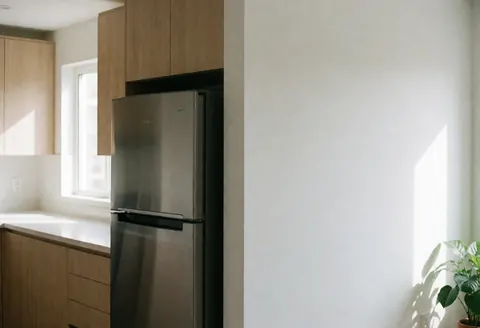 Un réfrigérateur en acier inoxydable se dresse dans une cuisine moderne avec des armoires en bois clair, des murs blancs et la lumière naturelle entrant par la fenêtre.