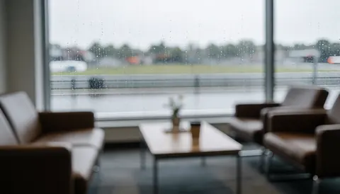 Intérieur d'une salle d'attente moderne équipée de canapés marron, d'une table basse et d'une grande fenêtre offrant une vue sur une piste de circuit automobile par temps pluvieux.