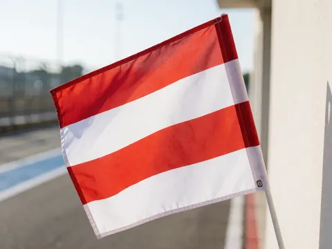 Détail d'un drapeau rouge et blanc signalant la voie des stands sur un circuit automobile, avec l'asphalt et les barrières floues en arrière-plan sous une lumière du jour vive.