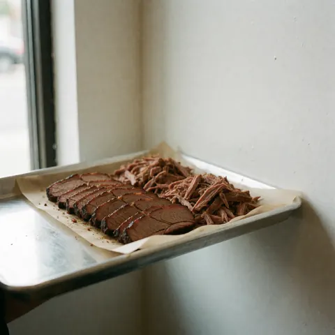 Assiette contenant de la viande grillée et de la viande effilochée posée sur du papier parchemin, le tout disposé sur un plateau métallique à côté d'une fenêtre baignée de lumière naturelle douce.