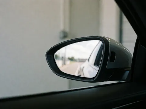 Détail du rétroviseur latéral d'un véhicule montrant le reflet d'une route, du ciel et de murs environnants dans une image réaliste et épurée.