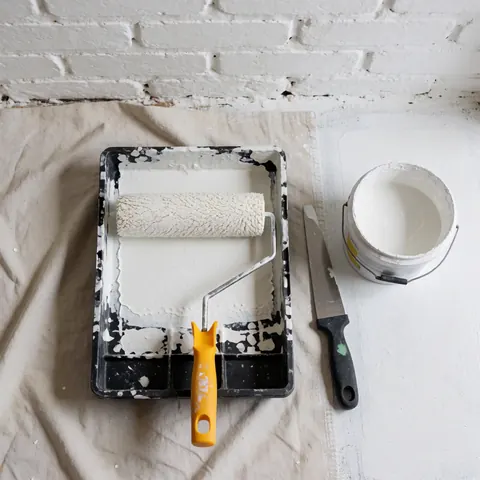 Rouleau de peinture blanc dans un bac, couteau de masquage et seau de peinture posés sur une toile de protection devant un mur de briques blanches.