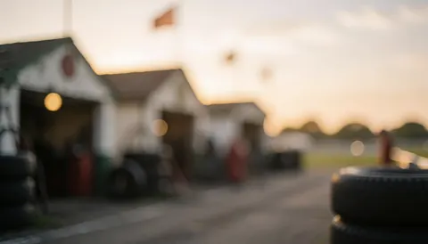 Vue floue d'une voie des stands de circuit avec des pneus empilés, des garages et une lumière dorée au coucher du soleil, servant de fond pour du texte commercial.