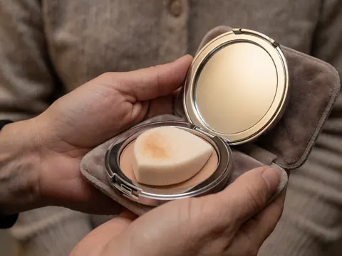 Éponge de maquillage utilisée posée à l'intérieur d'un miroir compact tenu par des mains, idéal pour les professionnels du maquillage.
