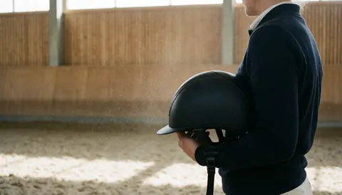 Un cavalier professionnel tient un casque d'équitation noir dans une vaste arène en sable avec des murs en bois et de la poussière en suspension.