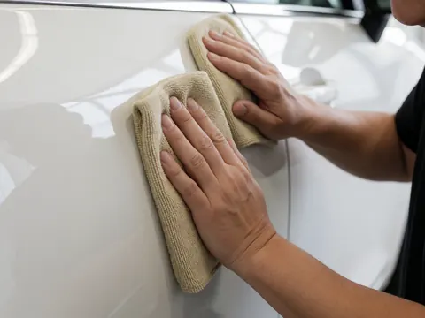 Main adulte tenant un chiffon microfibre pour polir la portière d'une voiture blanche brillante avec des reflets visibles sur un fond blanc uni.