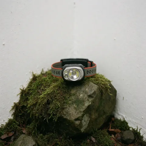 Une lampe frontale moderne repose sur un rocher recouvert de mousse humide, sur un fond blanc uni, évoquant la préparation pour des activités en plein air.